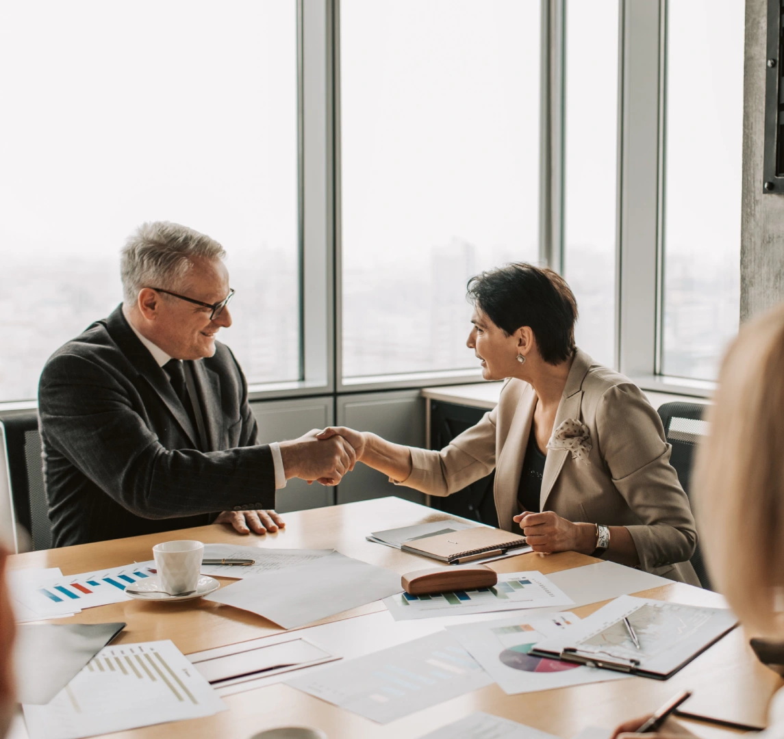 Two people in business attire shake hands over a conference table adorned with documents and graphs, embodying resilient leadership in a high-rise office setting.